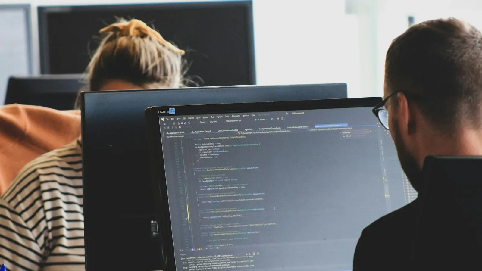 Two people working on computer code at monitors in a bright office workspace.