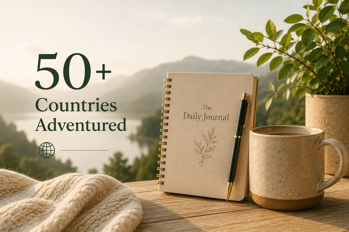 A beige spiral-bound daily journal titled 'The Daily Journal' sits on a wooden table with a black pen, next to a speckled ceramic mug and a knitted cream blanket, with a blurred mountain landscape and potted plant in the background. Text on the left reads '50+ Countries Adventured'.