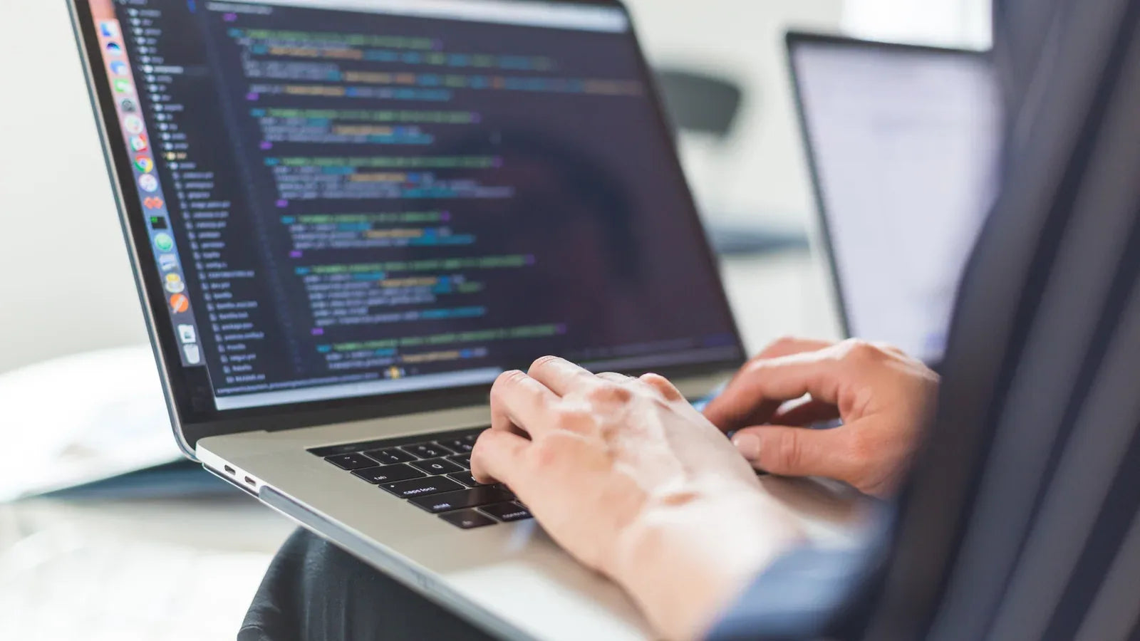 Developer typing code on a laptop beside a second screen.