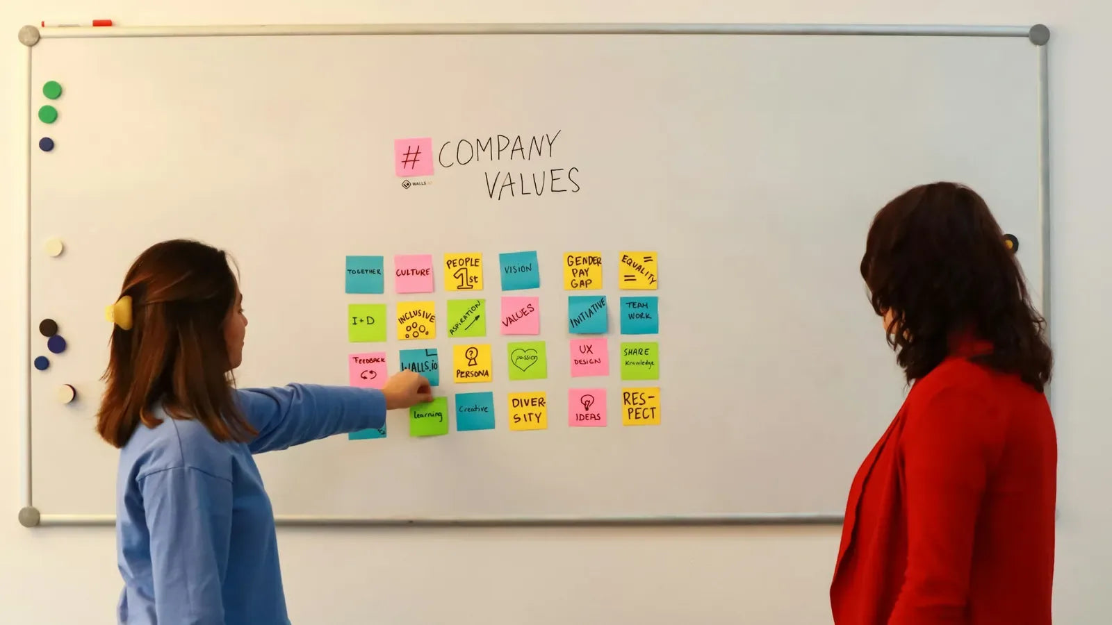 Two women standing in front of a white board with sticky notes on it.