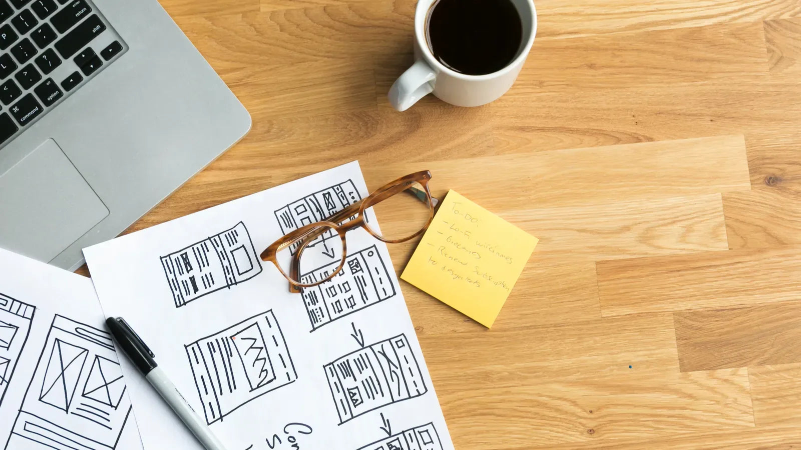 Hand-drawn website wireframes beside a laptop and coffee on a desk.